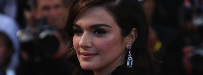 A woman with dark brown hair and wearing elegant earrings smiles softly while looking slightly to the side. The background is blurred but appears to show people and cameras, suggesting an event or gathering.