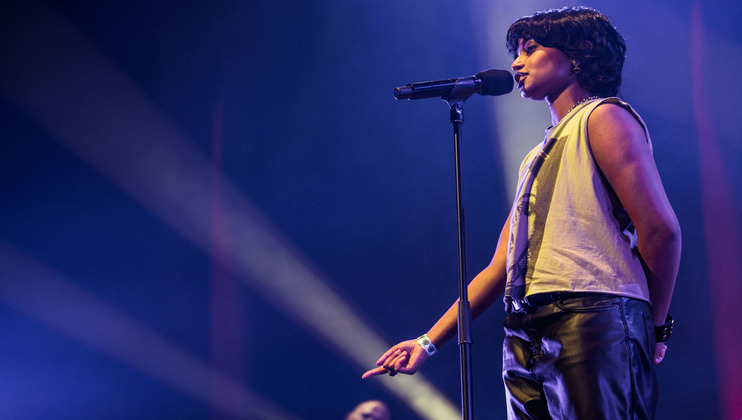 A singer stands on stage holding a microphone, illuminated by dramatic stage lighting. They are wearing a white sleeveless shirt and black pants, with a wristband on their left arm. The background is dimly lit with beams of light creating an atmospheric effect.
