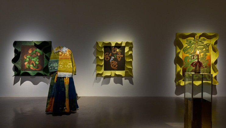 A gallery display featuring three wavy-framed artworks with vibrant colors. A mannequin dressed in a multicolored outfit stands in front of the left artwork. A glass showcase displays another item to the right of the middle artwork. Subdued lighting enhances the exhibit.
.