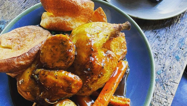 A plate of roasted chicken with a golden-brown skin, served with Yorkshire pudding, roasted potatoes, glazed carrots, and gravy. The plate is on a rustic wooden table with a second, partially visible dish in the background.
