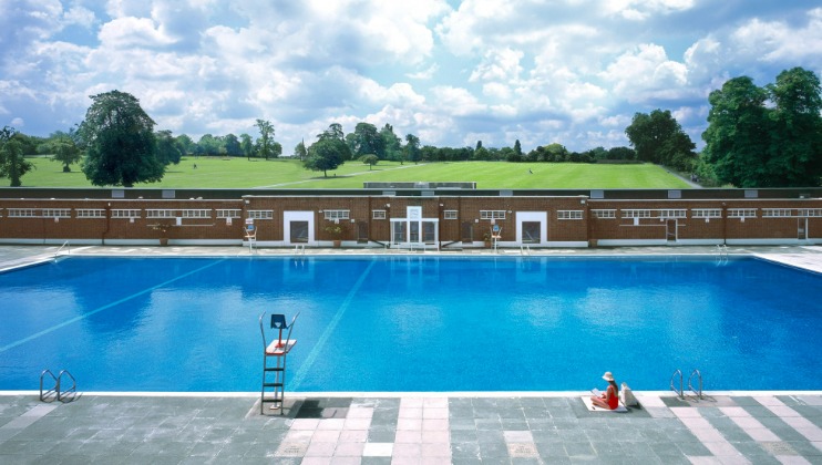 A large outdoor swimming pool with clear blue water is surrounded by a brick building. A lifeguard chair sits poolside, and a person in red attire is seated at the edge of the pool. Lush green hills and trees form the background under a partly cloudy sky.