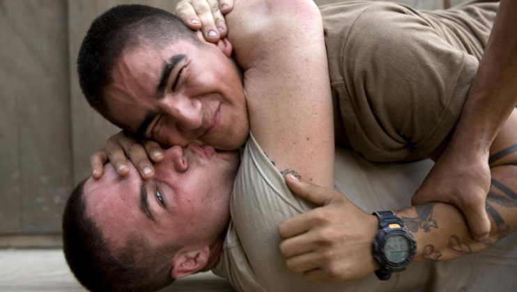 Two young men, both with short hair, are engaged in a playful wrestling match on the ground. One man, grinning, holds the other in a headlock while the other has his face scrunched up and his hand on the first man's face. Both appear to be in good spirits.