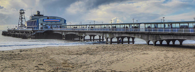 The image shows a pier extending from a sandy beach into the sea, with a building labeled Funfair Entertainments on it. The sky is cloudy, and the water appears calm. Numerous lampposts are lined along the pier.