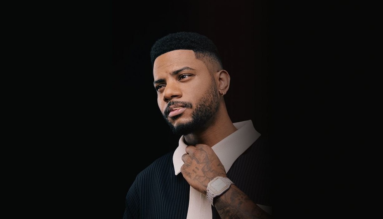 A man with a well-groomed beard and short hair, wearing a black and white collared shirt, poses against a dark background. He has a thoughtful expression, resting his right hand on his neck, and a wristwatch is visible on his left wrist.