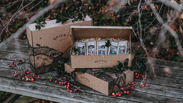 Cardboard boxes from Quicke's Traditional, a cheese company, are displayed on a wooden surface surrounded by greenery and berries. One box is open to reveal several cheese packages inside. The scene is set outdoors with twigs and leaves in the background.