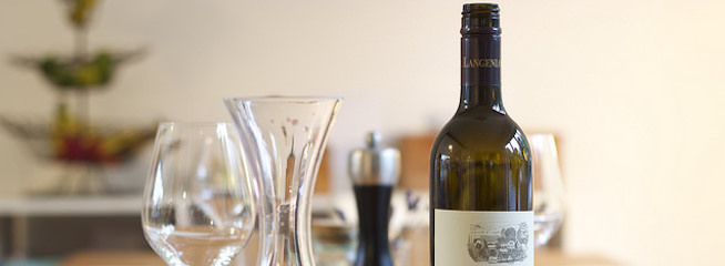 A bottle of wine with an elegant label is placed on a table set with empty wine glasses, a wine decanter, and a pepper grinder. The background is softly blurred, highlighting the main objects in the foreground.
