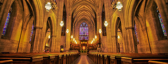 A grand interior of a cathedral with high, vaulted ceilings and intricate stained glass windows. Rows of wooden pews line the aisle leading to the altar, illuminated by warm hanging chandeliers. The atmosphere is serene and reverent.