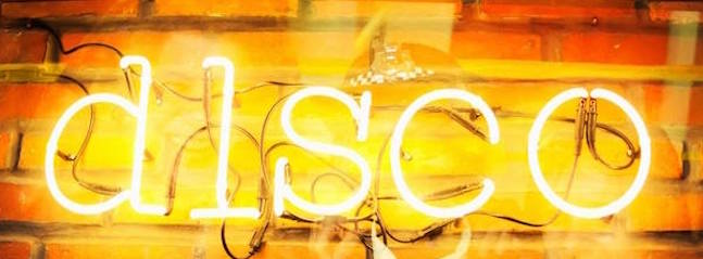 A neon sign with the letters d, i, s, c, o lit up in white against a brick wall background. The sign is slightly blurred, giving it a vibrant and dynamic appearance evocative of a lively atmosphere.