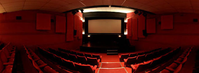 A dimly lit empty movie theater with red cushioned seats, a large projection screen at the front, and several ceiling lights illuminating the space. The theater hall has an intimate and cozy atmosphere with rows of seats gradually inclining towards the back.