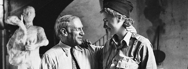 A black-and-white photo shows an older man wearing a shirt and tie with his arm around a smiling younger person dressed in military attire. They stand inside a room with a sculpture in the background.