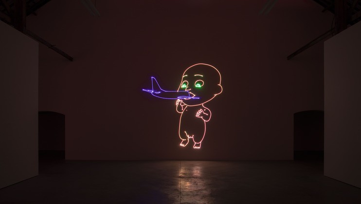 Neon artwork of a baby in an empty, dimly lit room. The baby is depicted with exaggerated, cartoon-like proportions and is shown holding a neon airplane close to its mouth. The neon lights are primarily blue, pink, and green.