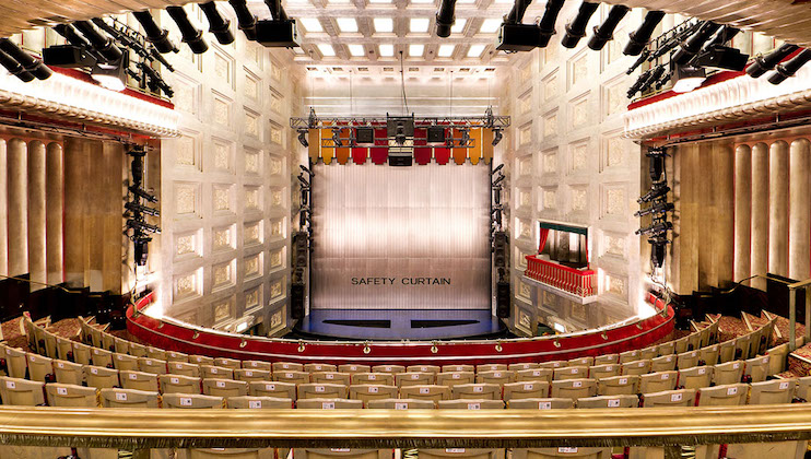 A spacious and elegant theater interior with red and white seats is shown from the back of the auditorium. The stage has a safety curtain that is lowered. The walls are adorned with ornate patterns and the ceiling features intricate lighting fixtures.