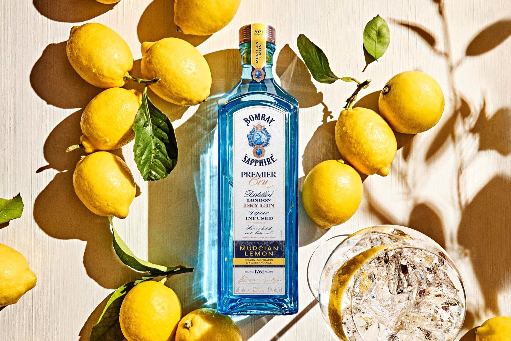 A blue bottle of Bombay Sapphire Premier Cru gin surrounded by fresh lemons on a light surface. The bottle is placed next to a glass filled with gin and ice, capturing bright sunlight and casting shadows.