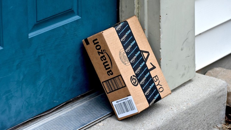A cardboard package with Amazon branding is placed on the doorstep of a blue front door. The box is sealed with black Amazon Prime tape and contains a barcoded shipping label. The doorstep is part of a grey concrete entryway with white house siding visible.