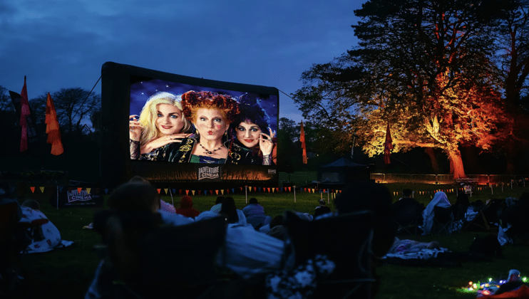 An outdoor cinema screening at dusk shows three women, likely from a movie, on a large inflatable screen. People are sitting on the grass, wrapped in blankets, watching the film. The background features trees illuminated by colorful lights and festively decorated banners.