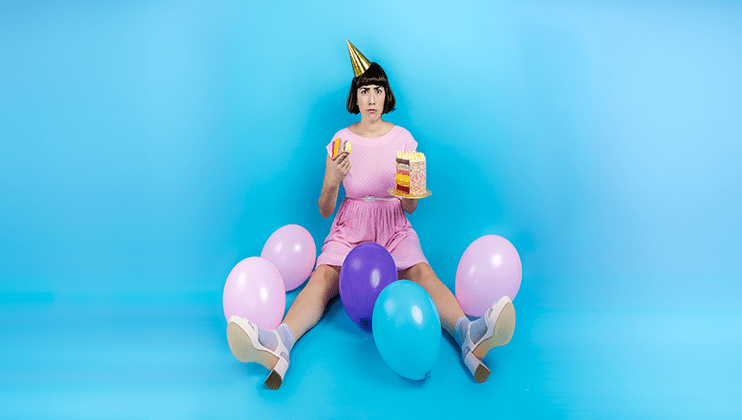 A person wearing a party hat and a pink outfit sits on the floor in a blue room, surrounded by colorful balloons. They are holding a slice of cake in one hand and a layered cake on a stand in the other, looking directly at the camera.