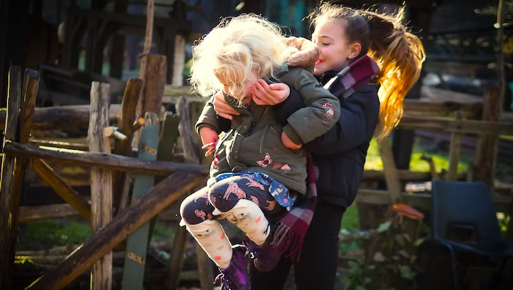 Two children are playing outdoors near a wooden fence. One child with blonde hair is being carried on the back of another child with long brown hair. Both wear winter jackets; the blonde child has colorful leggings showing animals while the other wears black pants.
