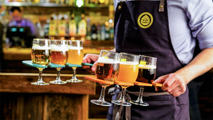 A server in a dark apron carries two trays filled with an assortment of beers in various glassware, from light to dark, in a bar setting. Bottles and glasses are visible in the background.