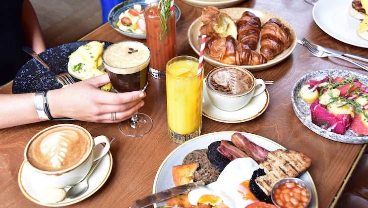 A table with a variety of breakfast foods, including eggs Benedict, croissants, fruit salad, and a full English breakfast. Drinks include coffee, orange juice, and a Bloody Mary. A person's hand holds a beverage on the left side of the image.