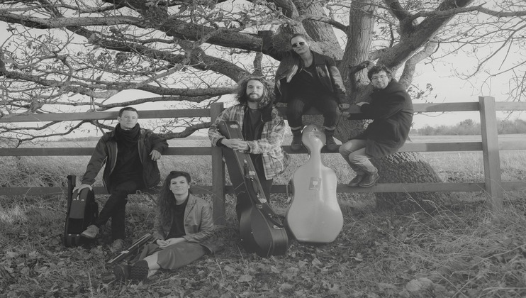 A black-and-white photo of five individuals in a field, posing next to a large tree and wooden fence. Three people are standing, two with guitars, and one with a cello case. Two others are sitting, one on the fence holding a violin case, and the other on the ground.