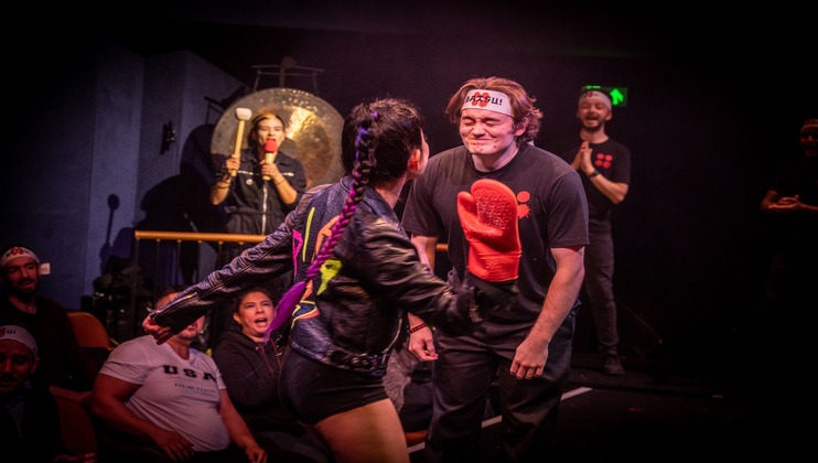 A person in a black jacket with colorful designs leans towards a smiling man wearing a headband and holding a large red mitten. In the background, a person strikes a gong while others look on, some appearing amused. The setting seems to be an indoor stage.