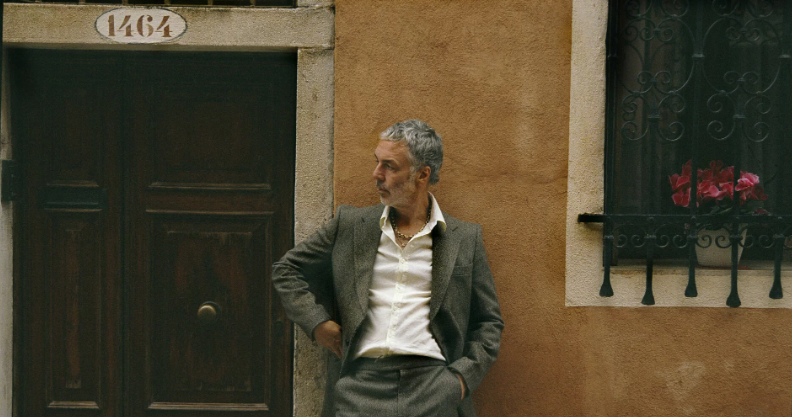 Baxter Dury stands againt a burnt sienna wall beside a mahogany door and an intricate iron window guard behind which sit vivid pink flowers. He looks to the left with a hand in his grey suit pocket.