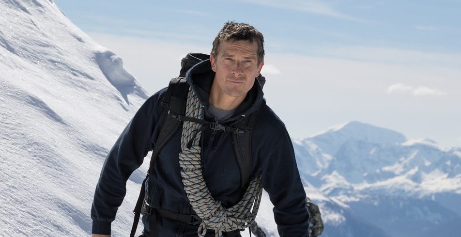 A man dressed in outdoor gear with a climbing rope slung over his shoulders is standing on a snowy mountain slope. He has a backpack and is looking forward. In the background, there are snow-covered mountains under a blue sky.