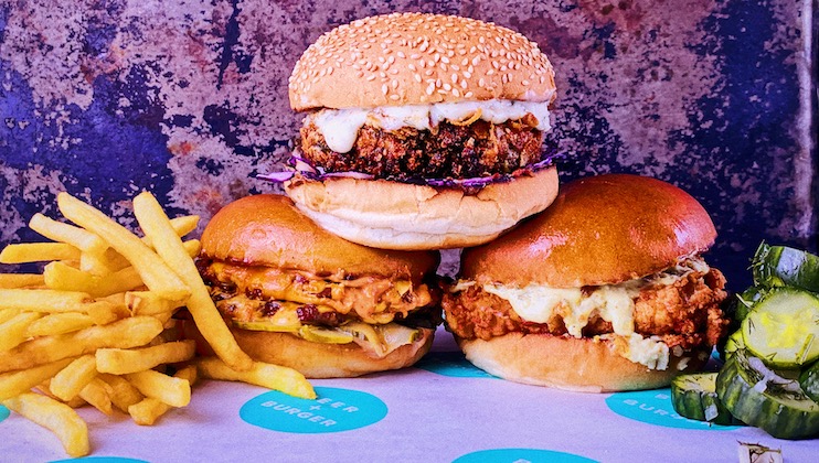 Three savory hamburgers are displayed along with a side of crispy French fries and slices of pickles. The burgers feature an assortment of toppings including melted cheese and fresh lettuce, all set on a turquoise and white patterned table cover with Sister Burger logos.