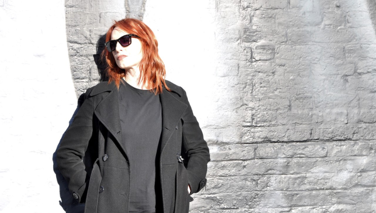Musician Beth Rettig, in a black coat and black shades, stands against a grey wall
