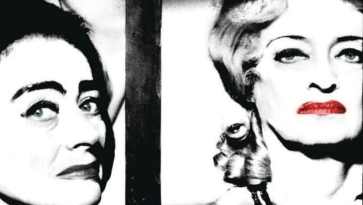 Black and white photo of two women with intense expressions. The woman on the left has dark eyebrows and the woman on the right has lighter, curled hair and bold red lipstick. Both appear to be looking slightly off-camera, evoking a dramatic mood.