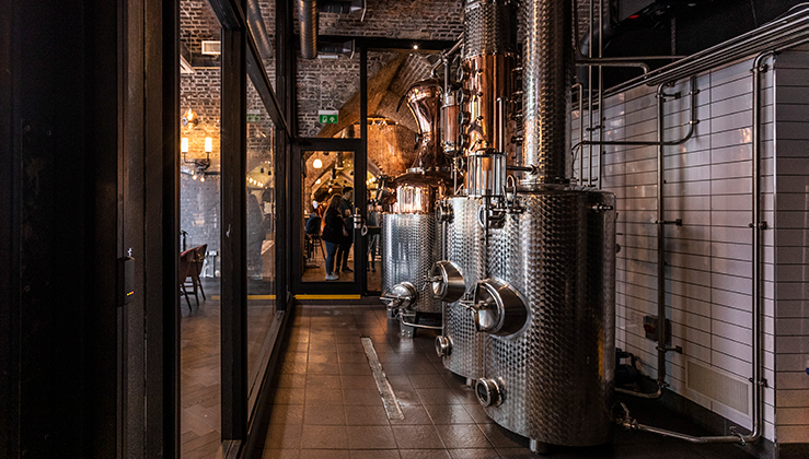 A distillery with shiny stainless steel industrial equipment. The room has a brick ceiling and one glass wall, through which other parts of the distillery can be seen. In the distance, a person appears to be walking towards the equipment.