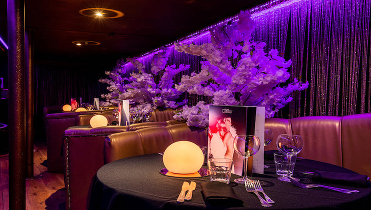 A dimly lit, elegantly set dining area with round tables adorned with glowing crystal ball lamps, neatly placed glassware, and menus. Faux cherry blossom trees and beaded curtains, all under purple lighting, create a luxurious and intimate ambiance.