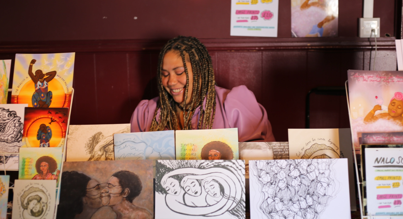 A smiling artist sits at her stall at Black Brighton Market, selling prints.