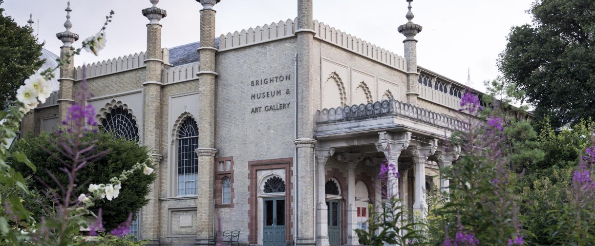 The image shows the exterior of the Brighton Museum & Art Gallery, a historical building with ornate architectural features, including columns and arched windows. There are green trees and blooming flowers in the foreground, adding color to the scene.