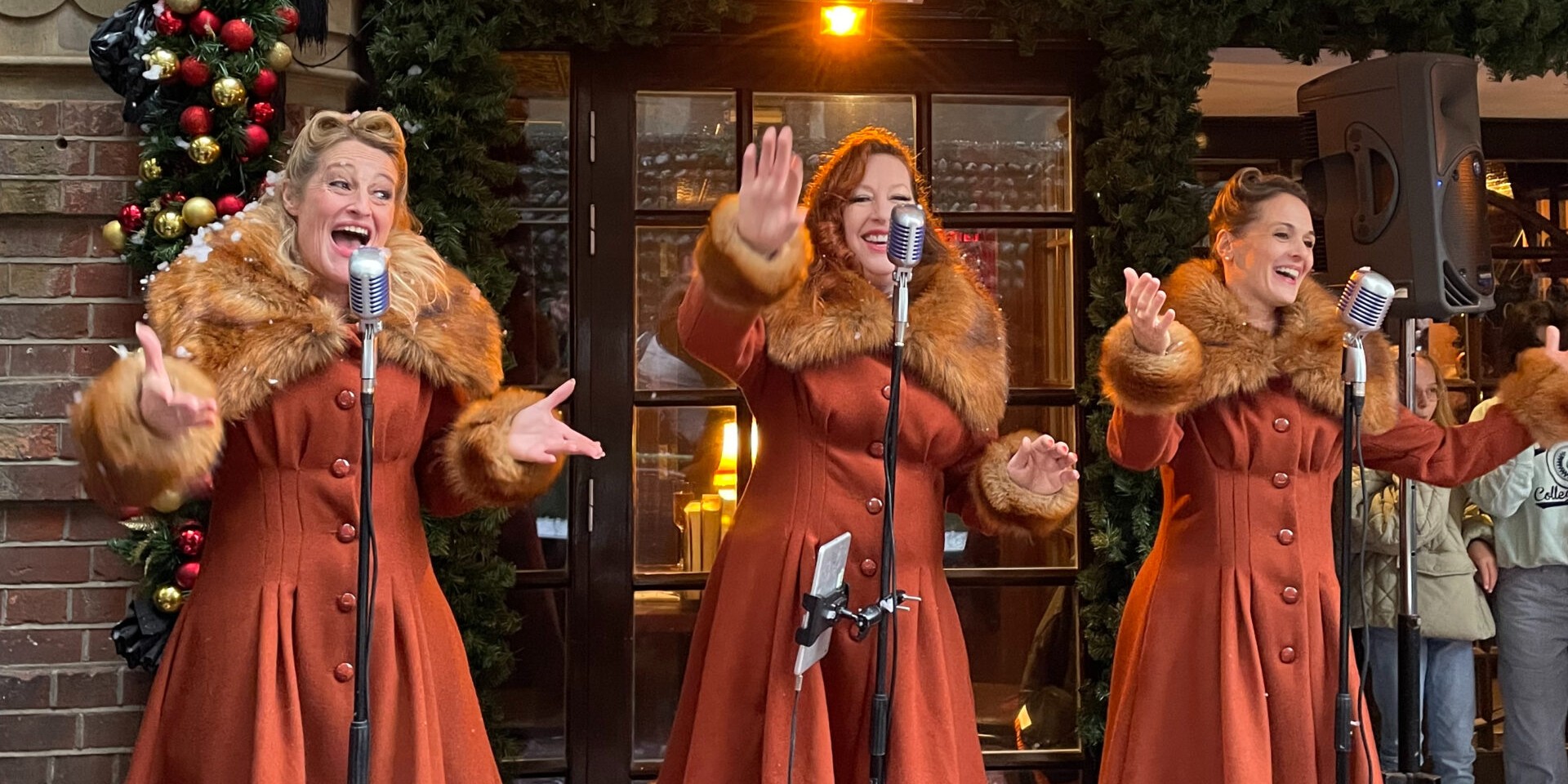 Three joyful singers dressed in lush fur coats perform on the street in front of Christmas wreath bunting.