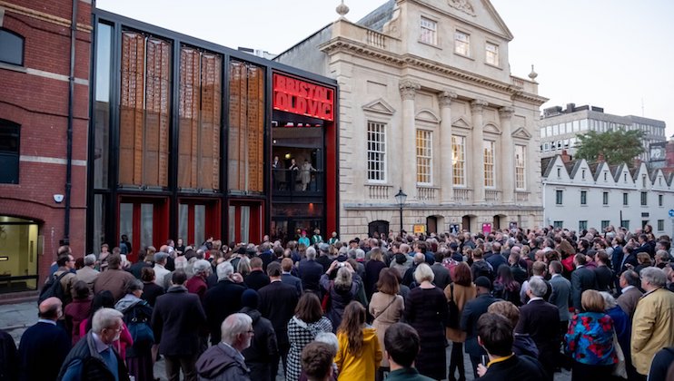 A large crowd gathers in front of the Bristol Old Vic theatre. The building features a modern glass facade with a prominent red sign that reads Bristol Old Vic, alongside a classical building with large columns and arched windows. The atmosphere is lively.