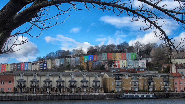 A row of colorful houses sits on a hillside covered with bare trees. Below the hill are two-story buildings with balconies facing a waterway, where a black narrowboat is moored. A large tree branch frames the image from the top. The sky is partly cloudy.