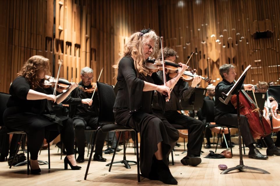 A group of musicians in formal black attire perform on stage with string instruments, including violins and a cello. They are seated in front of a wooden panel backdrop, concentrating on their playing. The stage lighting creates a warm ambiance.