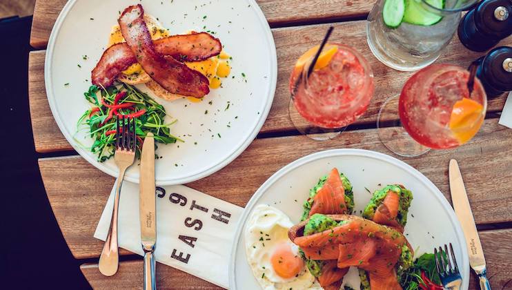 A wooden table with two plates of gourmet food. One plate features bacon over eggs and salad greens, while the other has smoked salmon, avocados, and poached eggs. Two glasses of pink drinks with orange slices and a jug of water with cucumber slices are also on the table.