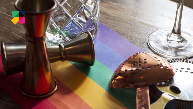 A variety of cocktail making tools, including a jigger and strainer, are placed on a rainbow-striped surface. A glasses and a crystal jar are also present. In the top left corner, a colorful geometric logo is visible.