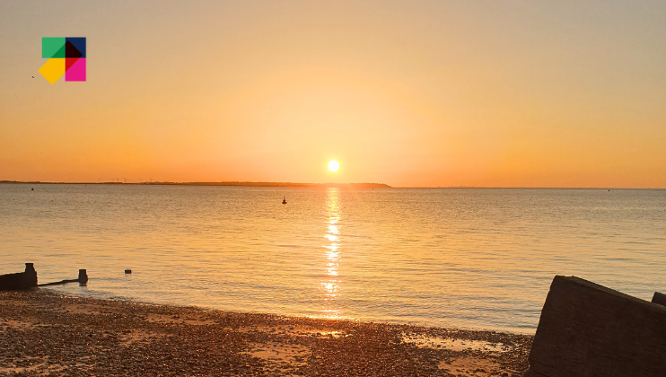 A serene sunset over a calm beach with pebbles on the shore. The sun casts a golden reflection on the tranquil water, and a clear sky adds a warm orange and yellow hue. A colorful, geometric logo is positioned in the upper left corner of the image.