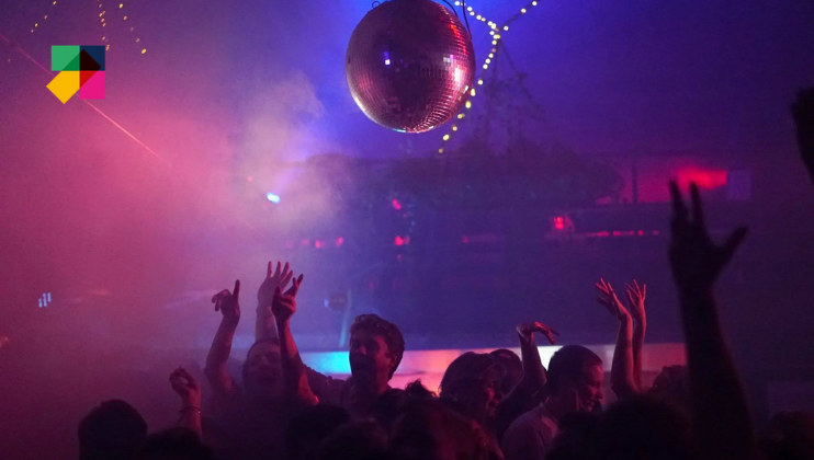 A crowded dance floor with colorful lights, a shiny disco ball overhead, and fog creating a hazy atmosphere. People are raising their hands and dancing, immersed in the vibrant party scene. A multicolored logo is displayed in the top left corner.
