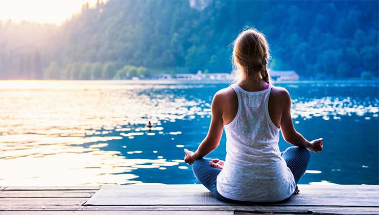 A person with long hair sits cross-legged on a wooden dock, meditating by a serene lake surrounded by lush, green hills. The individual is facing the water, wearing a white tank top and patterned leggings, with a tranquil and peaceful ambiance.