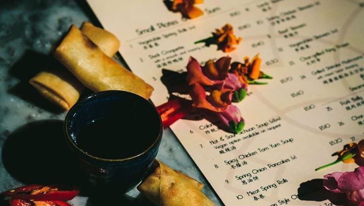 A menu page with various food items written on it, decorated with vibrant flower petals and accompanied by two spring rolls and a ceramic cup placed on a textured surface. The setup is in a lightly dimmed, possibly rustic, environment.