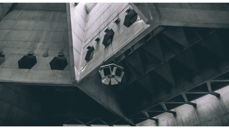 A black and white image of an architectural interior featuring modern, geometric concrete designs. The ceiling has triangular and polygonal shapes with a central suspended light fixture. The sharp angles and clean lines give a minimalist, futuristic feel.