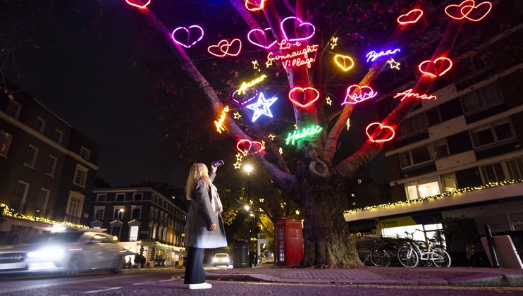 A woman stands on a street at night, gazing at a large tree adorned with vibrant, multicolored neon lights shaped like hearts, stars, and words such as Peace, Love, and Hope. The surrounding street and buildings are also illuminated with festive lights.
