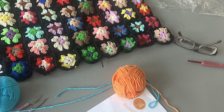 A desktop with the corner of a crotchet granny square blanket, two balls of wool, some reading glasses, scissors and a crotchet needle.