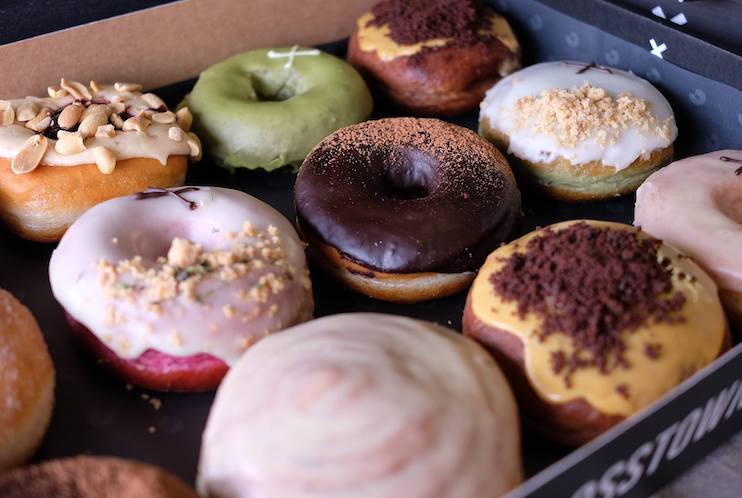A close-up of an assorted dozen donuts in a box. The donuts have various toppings and glazes, including green matcha with nuts, chocolate glaze with cocoa powder, white glaze with sprinkle toppings, and a pink glazed donut.