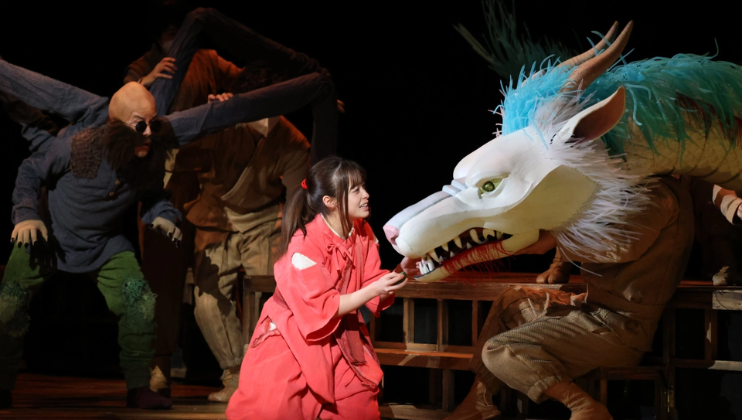 A performer in a red costume interacts tenderly with an actor in a dragon costume on stage. Surrounding them are other cast members dressed in various theatrical outfits, contributing to an elaborate scene. The dragon costume is detailed with a blue mane and white fur.