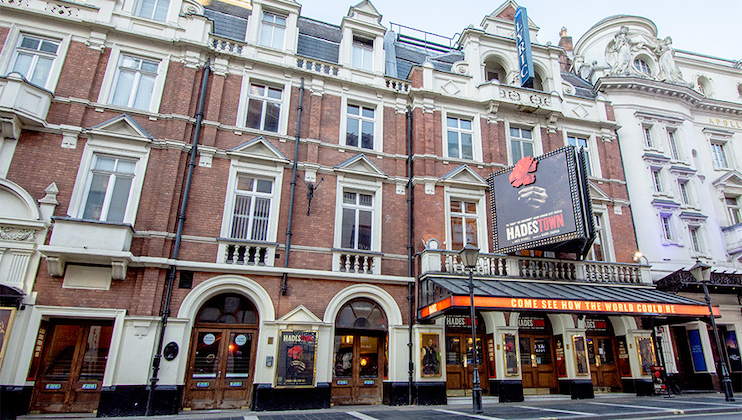 A historic brick theater building with arched windows and ornate architectural details. A marquee displays Hadestown, and a large promotional poster for the show is hanging on the facade. The entrance is illuminated, inviting patrons inside.